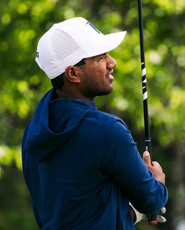 Man in blue jacket and white cap holding a golf club with greenery in the background