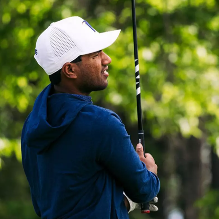 Man in blue jacket and white cap holding a golf club with greenery in the background